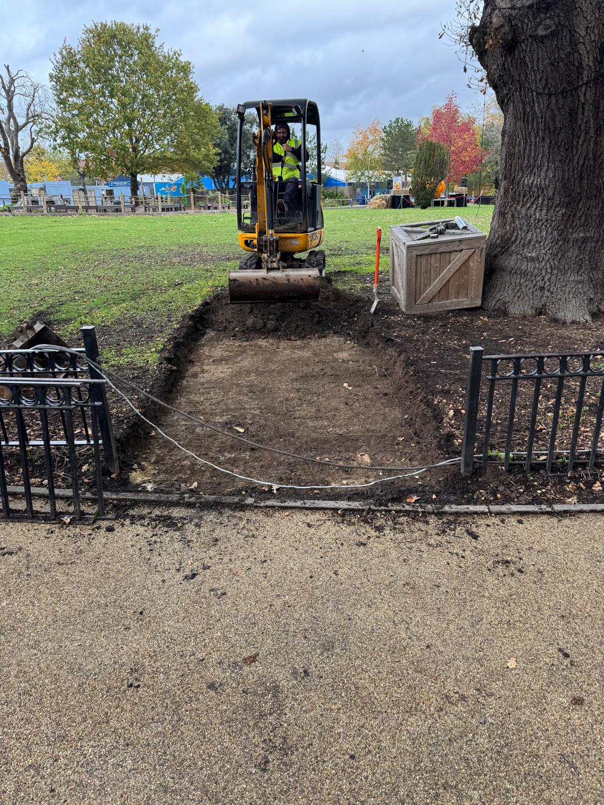 Mini digger excavating a footpath formation through grass beside a mature tree and iron railings
