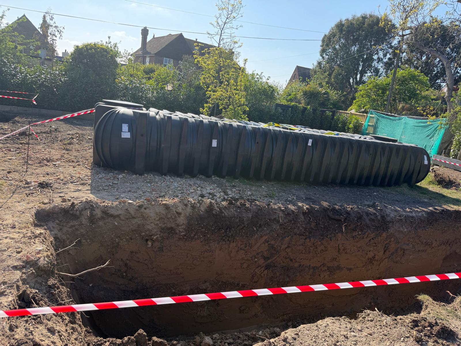 Large underground rainwater storage tank staged next to a square excavation with safety barrier tape
